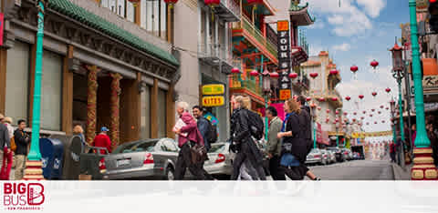 This image shows pedestrians crossing a vibrant street with traditional Chinese architecture and hanging red lanterns. A clear sky is above and a vehicle is visible. The Big Bus Tours logo appears in the corner, suggesting a sightseeing tour location.