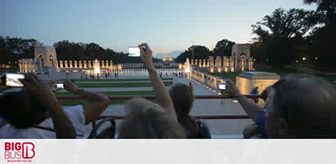 Visitors on a Big Bus tour observe a well-lit, reflective pool and monuments at dusk from the bus’s upper deck. The foreground shows the back of people’s heads, some with arms raised to take photos. Trees line the horizon under a fading sky.