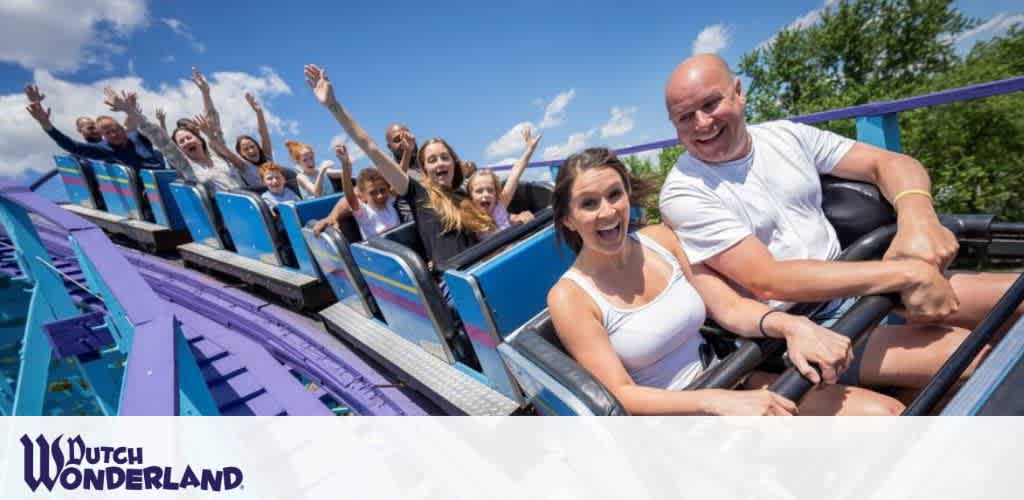 Excited visitors on a roller coaster at Dutch Wonderland. They are smiling and raising their hands in the air under a clear blue sky, as the coaster descends a vibrant blue track. The park's logo is visible in the bottom left corner.