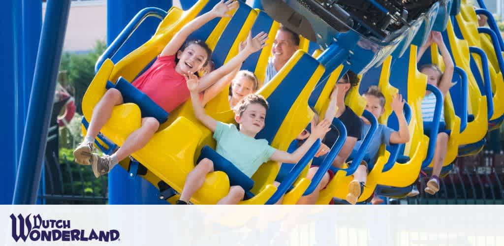 Image shows an exhilarating moment on a roller coaster with children and adults. They are seated securely in a row on a blue and yellow ride, arms raised, with expressions of joy and excitement. The backdrop features a clear sky and part of the attractions structure. Logo of Dutch Wonderland is visible in the lower left corner.