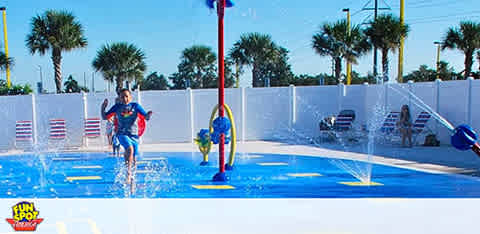 Child plays in a splash pad area with water jets on a sunny day.