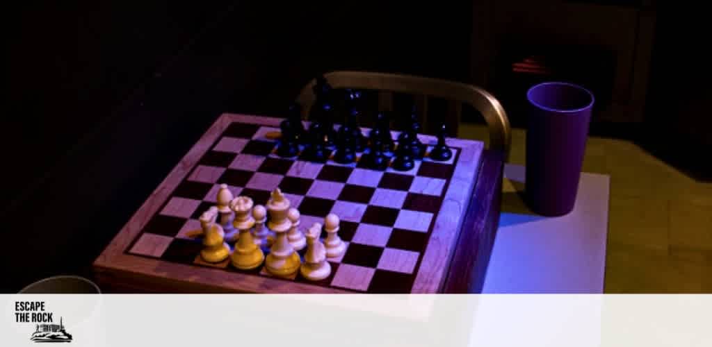 This image features a close-up view of a chessboard with a complete set of wooden chess pieces arranged and ready for a game. The chessboard is placed on a table with a brown chair partially visible on one side. The surface of the table and the chair have a reflective sheen, suggesting a smooth material. There is a purple mug to the right of the chessboard. The lighting casts a dramatic atmosphere with shadows and a predominantly dark background, highlighting the chess pieces and giving a focus on the strategic game setup. The logo "ESCAPE THE ROCK" is visible in the lower-left corner indicating an escape room or puzzle game theme. The elements of the image may suggest an inviting challenge or intellectual pastime.

At FunEx.com, we're proud to offer the opportunity to exercise your mind at the lowest prices — be sure to take advantage of our exclusive discounts on tickets for a variety of intriguing and immersive experiences!