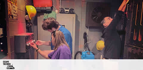 Image Description: This is a vibrant photograph taken in an escape room, showcasing a group of three participants engaged in solving a puzzle. On the left, we see an individual with mid-length hair and a blue top, concentrating on a device in their hands, looking for clues. The person in the center, appearing in a purple shirt, is closely examining another component of the game, perhaps a lock or a code device. On the right, a third person is reaching up towards what appears to be a set of shelves or compartments, searching for potential hints or items that could aid in their escape. The room is dimly lit to enhance the mysterious atmosphere, with practical props and yellow balloons adding to the thematic environment. The phrase "ESCAPE THE ROCK" is visible on the wall, indicating the theme or name of the escape room challenge.

Experience the thrill of an escape room adventure and get your tickets at FunEx.com, where you can unlock savings with the lowest prices available.