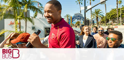 A tour guide smiling with a microphone on a sunny day with passengers on a sightseeing bus.