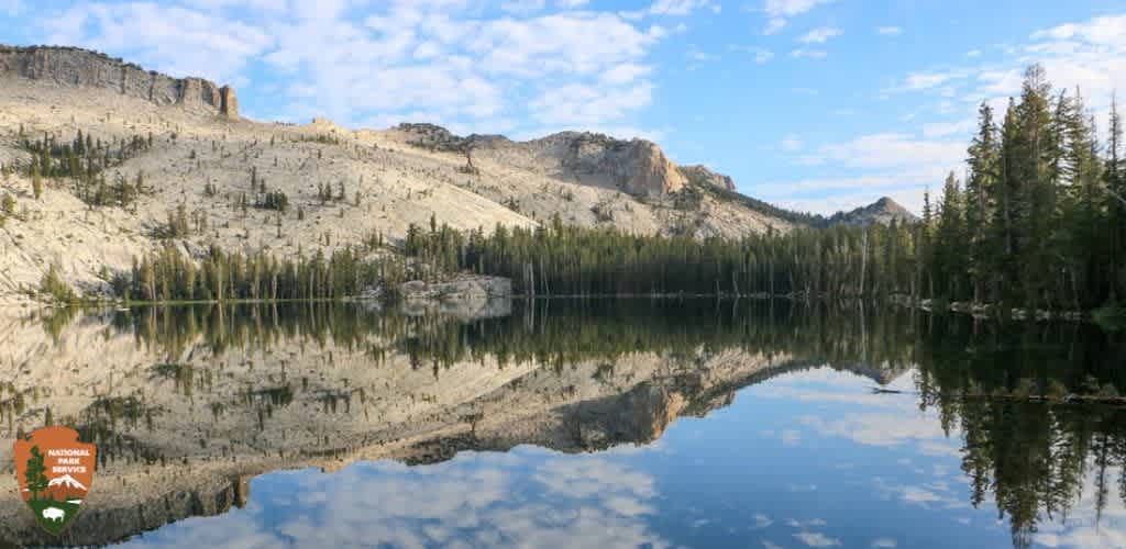 This image presents a serene view of a mountainous landscape reflected in the still waters of a calm lake. The sky is partly cloudy, allowing for portions of blue to peek through and mirror off the water's surface. On either side of the lake, there are forested slopes leading up to rugged cliffs and peaks, which show a variety of vegetation, including coniferous trees and scattered patches of bare rock or stone. The trees and sky create a symmetrical reflection in the water, providing a sense of tranquility and unspoiled nature. In the lower left corner of the image, there's a logo for the National Parks Service suggesting that this scenic view could be located within one of the national parks.

Don’t miss out on the breathtaking beauty nature has to offer; visit FunEx.com to find exclusive discounts and the lowest prices on tickets for your next adventure.