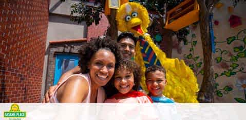 A joyful family poses with a large, yellow Big Bird character at Sesame Place. Two children and their smiling parents are in the foreground with the costumed character behind, set against a colorful backdrop with red brick and greenery. The Sesame Place logo is visible in the corner.