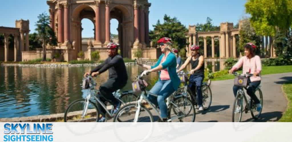 Four cyclists with helmets ride past a grand structure by a waterway under a clear sky.