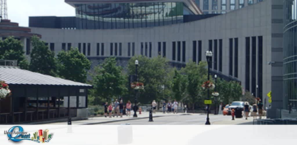 Image shows a sunny day in an urban plaza with pedestrians walking in the distance. A vehicular street crosses the frame, and modern buildings with large windows are seen in the background. A small structure adorned with hanging flower baskets is visible on the left. The logo for Urban Adventure Quest partially overlays the image at the bottom.