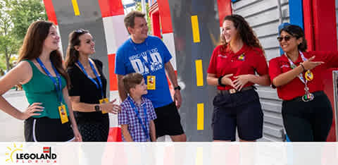 Visitors enjoy an interaction at LEGOLAND. Two staff members in red uniforms engage with a group of two women and a man, all holding ID badges, accompanied by a young boy. They stand in front of a colorful LEGO-inspired structure, indicating a playful and welcoming theme park atmosphere.