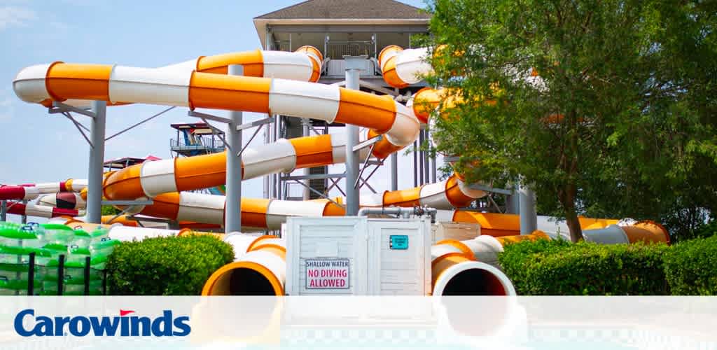 Image depicts a vibrant water park scene centered on a large, twisting water slide with orange and white color scheme. The slide structure emerges from a verdant landscape under a clear sky. The foreground shows a clear, calm pool with a sign that reads 'Shallow Water No Diving Allowed.' The watermark 'Carowinds' is visible at the bottom.