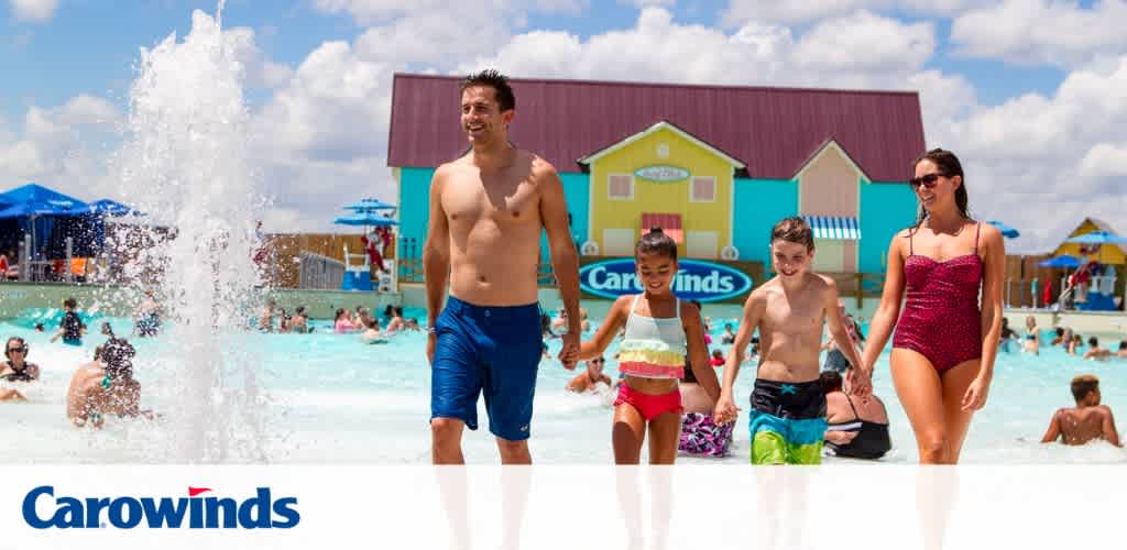 Image of a sunny waterpark with clear blue skies. In the foreground, a man walks alongside two children, holding hands with the girl to his right. A woman accompanies them, all wearing swimwear. Behind them, visitors enjoy the water, and colorful buildings line the backdrop.  Carowinds  logo visible at bottom.