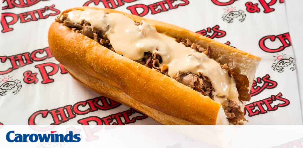 Image features a close-up of a cheesesteak on a crisp roll, filled with sliced steak and melted cheese. The background shows a patterned paper with 'Chicken Pete's' text and logo. The word 'Carowinds' is visible in the foreground, indicating the location.