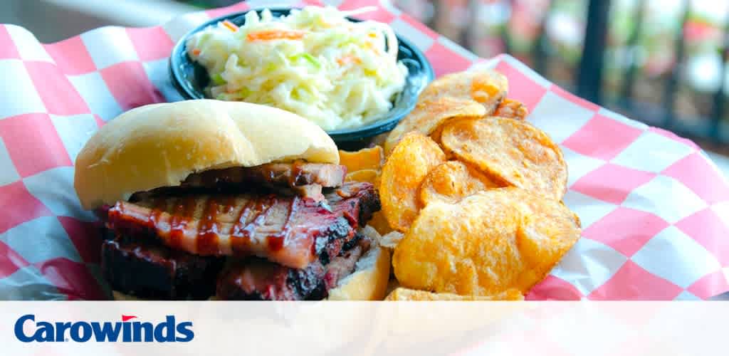 Image shows a barbecue sandwich with slices of brisket on a bun, paired with a side of coleslaw and seasoned potato chips. The meal is presented on a paper-lined tray with a pink and white checkered pattern. The background is softly blurred, and the logo of Carowinds is visible in the lower portion of the image.