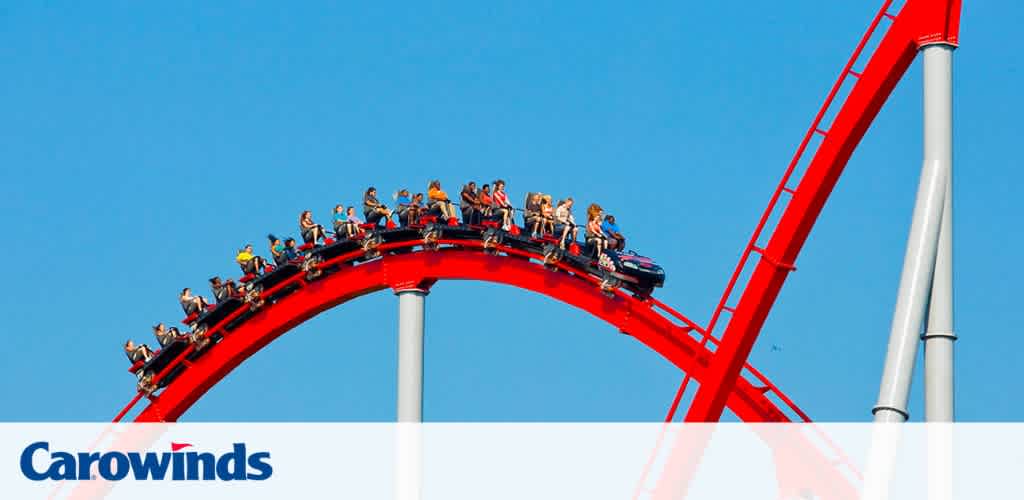 Image shows a red roller coaster with a train of passengers cresting a curve against a blue sky. Riders are seated in rows with over-the-shoulder restraints, appearing excited. Below, the Carowinds logo is visible, indicating the location is Carowinds amusement park.