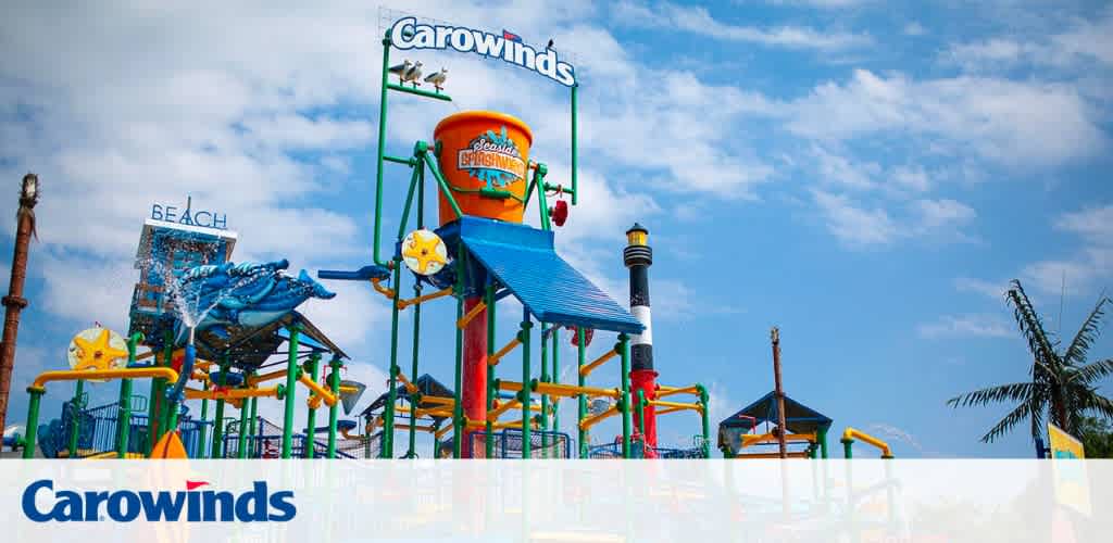 Image of a colorful water play structure at Carowinds amusement park under a blue sky. Features slides, interactive water elements, and a large tipping bucket atop. The park's name and logo are prominently displayed.