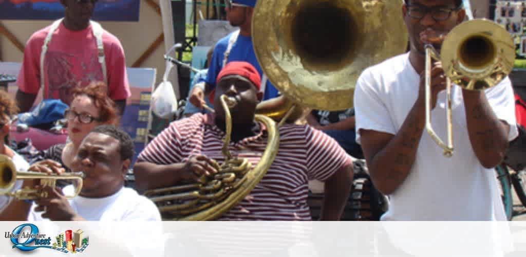 An outdoor scene with three musicians playing brass instruments, a crowd behind them. The first is seated with a large tuba, the second stands holding a sousaphone, and the third plays a trumpet. Sunlight bathes the group in a cheerful ambiance.
