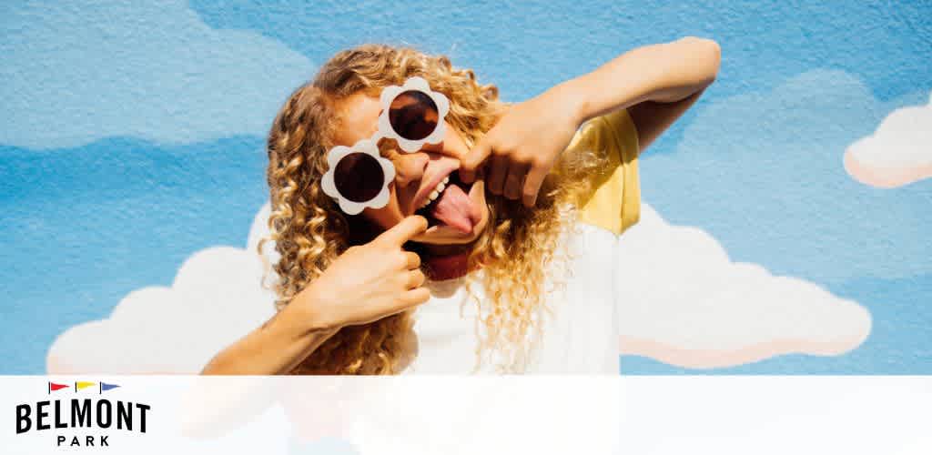 Image description:

This is a vibrant and playful promotional image featuring a woman against a blue sky with white clouds painted on a wall. The woman has curly blonde hair and is wearing large, round sunglasses that resemble smiling faces with their top halves being white and the lower halves dark, creating the illusion of eyes. She is playfully sticking her tongue out to the side in a cheeky gesture and is leaning on a white horizontal barrier that separates her from the illustrated sky. The lower left corner contains the logo for "Belmont Park" with a colorful design above the text.

Visit FunEx.com and let the fun soar with incredible discounts and savings on tickets - get ready to experience the thrills at Belmont Park with the lowest prices available!