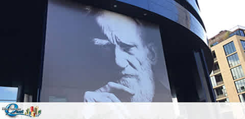 An outdoor billboard features a large black and white close-up image of a contemplative individual with facial hair resting their chin on their hand. The surrounding area appears to be urban, with parts of buildings visible under a clear sky. The billboard is a part of a curved structure.