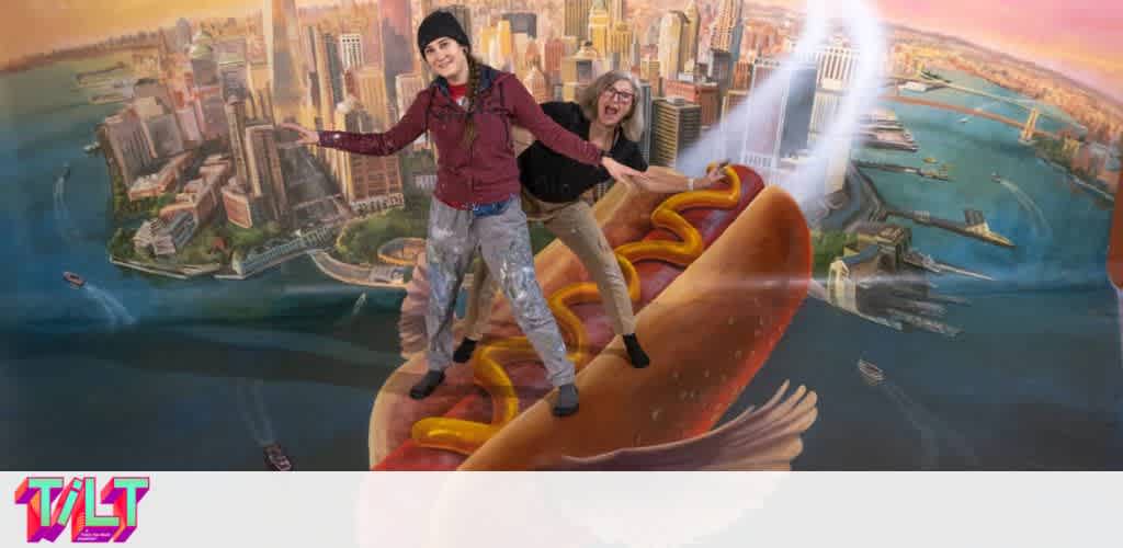 Two people are posing with exaggerated expressions as if surfing on a hotdog soaring above a cityscape, with skyscrapers and waterways below. The background mural is designed to create an illusion of flying. The image includes the word  TILT  in multicolored text.