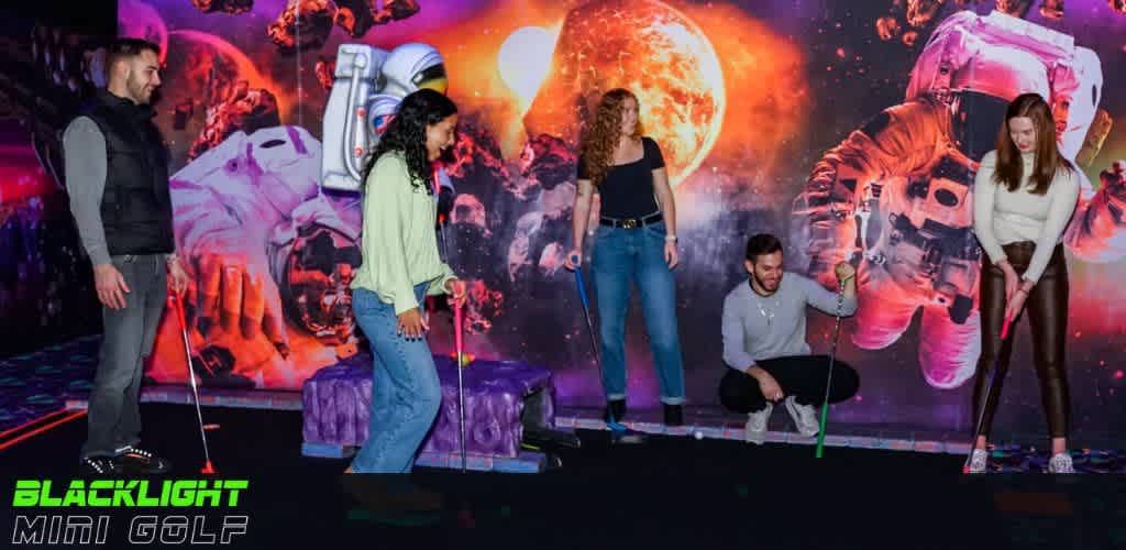 Four people are enjoying a game of blacklight mini golf, surrounded by vibrant space-themed murals with celestial bodies and astronauts. Two men and two women, each holding a putter, appear engaged and playful on the fluorescently lit course, enhancing the cosmic experience.