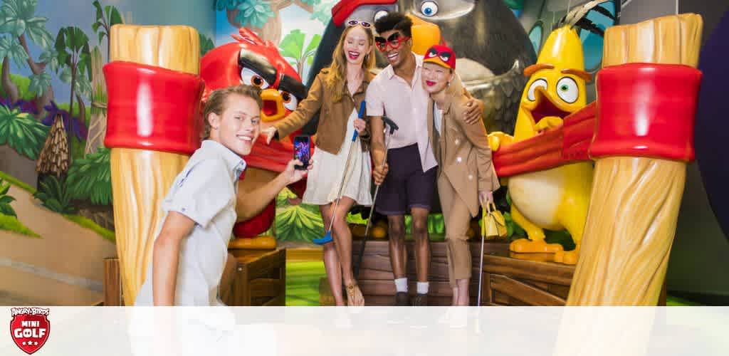 This image shows a joyous group at an Angry Birds mini-golf course. A young person in the foreground takes a selfie, while three others pose with clubs, smiling broadly amidst vibrant, oversized Angry Birds characters and tropical scenery.