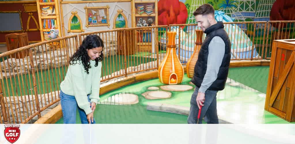 A woman and a man enjoy a game of indoor mini golf. The woman, focused, prepares to putt on the green course, while the man watches with a smile. Bright cartoonish game-themed murals decorate the walls, enhancing the playful atmosphere.