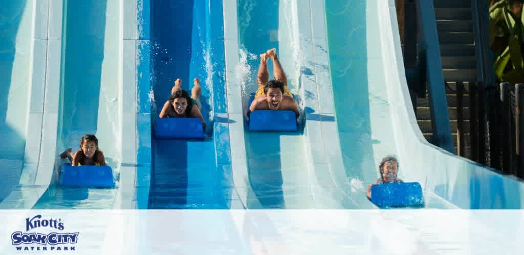 This image captures the excitement at Knott's Soak City Waterpark. It shows four individuals on adjacent blue water slides, each riding a dark blue mat. They are in various stages of descent, with water splashing around them, indicating the thrilling speed of their ride. The person furthest to the left is sitting up, perhaps mid-slide, while the others are lying on their backs, arms raised in excitement or to maintain balance. The bright sunlight casts a cheerful glow on the scene, enhancing the vivid blue of the slides. The Knott's Soak City logo prominently displayed at the bottom of the image reinforces the location.

For those looking to make a splash and save, FunEx.com offers the best discounts on Knott's Soak City tickets, guaranteeing you the lowest prices for your next aquatic adventure.