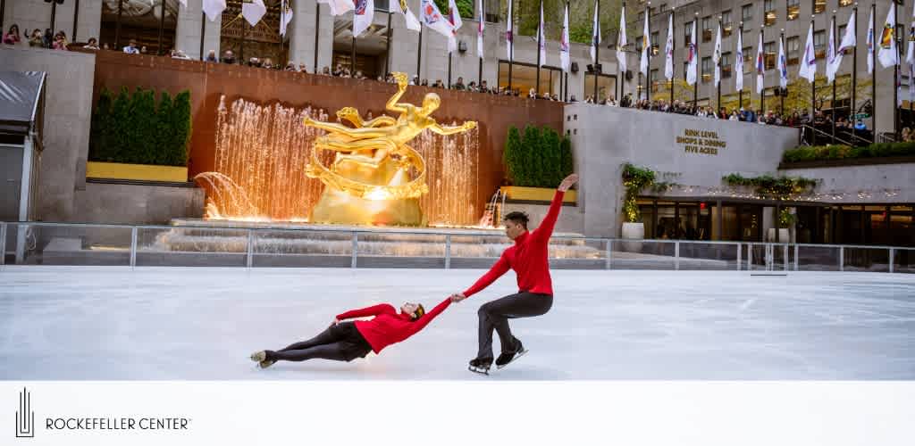 Two ice skaters perform at the Rockefeller Center rink. A person in red is spun low across the ice by their partner, creating a dynamic visual. They’re surrounded by flags, a golden statue, and observers above.