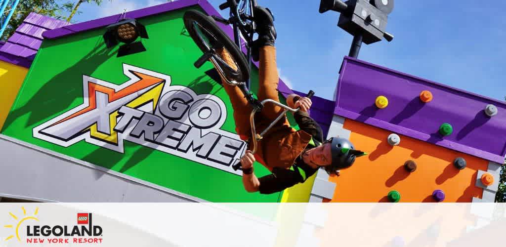 Image of an exhilarating moment at LEGOLAND New York Resort featuring a person in mid-air performing an acrobatic BMX trick against the vibrant backdrop of a 'GO XTREME' sign, with colorful Lego-style knobs adorning the structure. The sunny sky adds to the lively atmosphere of the scene.