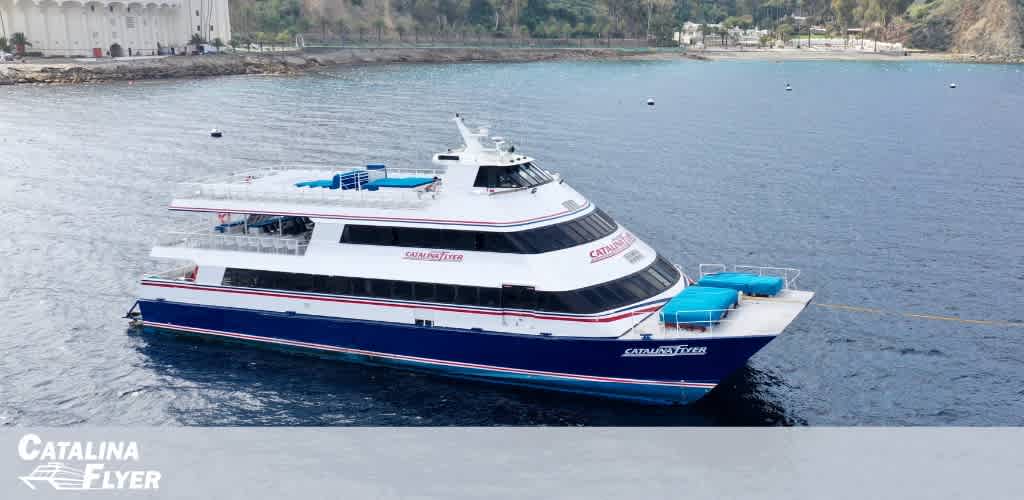 Image shows the Catalina Flyer, a large white, blue, and red passenger ferry, mid-voyage on a calm sea. The ferry's name is visible on its side, with serene water and soft hills in the backdrop. The Catalina Flyer logo is at the bottom of the photo.