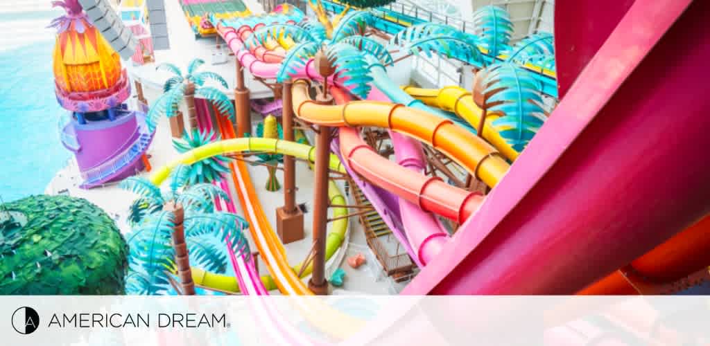 An elevated view of a vibrant water park with multiple intertwined water slides in bright colors of pink, yellow, and blue. Faux palm trees add to the tropical feel. The American Dream logo is visible on the bottom left.