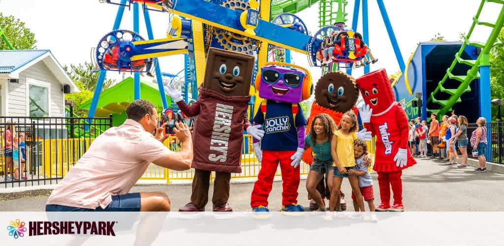At Hersheypark, a person kneels to take a photo of animated Hershey’s chocolate characters greeting enthusiastic children. The park's vibrant rides form a colorful backdrop under a clear sky.
