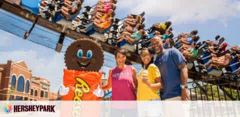 In the foreground, three smiling individuals stand next to a mascot resembling a chocolate bar, in front of a railing with a theme park logo. Behind them, riders on a roller coaster with their arms raised appear mid-descent, conveying excitement. Clear skies overhead.