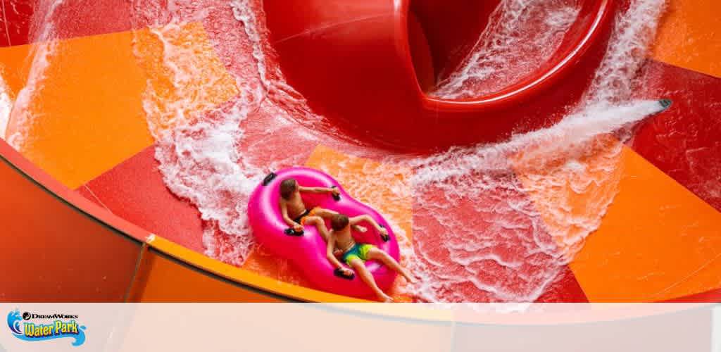 Image shows a vibrant water slide with a bright orange and red color scheme. Rushing water is channeled down the slide, and a person on a pink inflatable ring is enjoying the ride. The DreamWorks Water Park logo is visible in the corner.