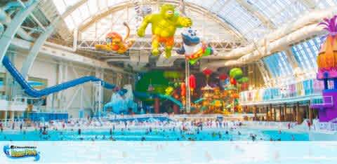 Indoor water park with a vibrant atmosphere, featuring a large, bustling swimming pool. Colorful, oversized inflatable character decorations hang from the ceiling. A blue water slide winds from the left, while the bright interior includes an array of balloons and lively architecture, giving a festive feel.