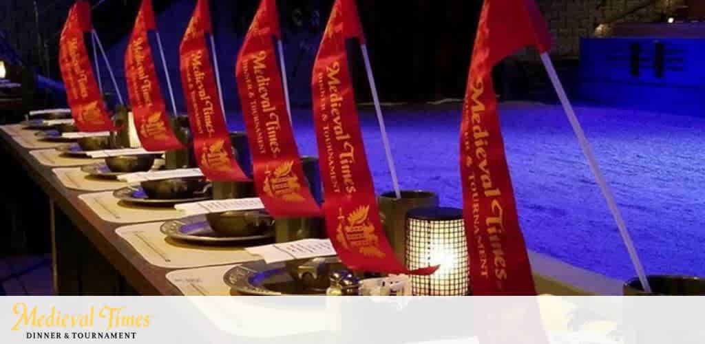 Image of a row of banquet tables set up for the Medieval Times dinner and tournament. Each place setting includes a yellow flag with the Medieval Times logo, red napkins, metal bowls, and drinking vessels. The arena background is dimly lit with a blue hue. The Medieval Times logo is visible in the foreground.