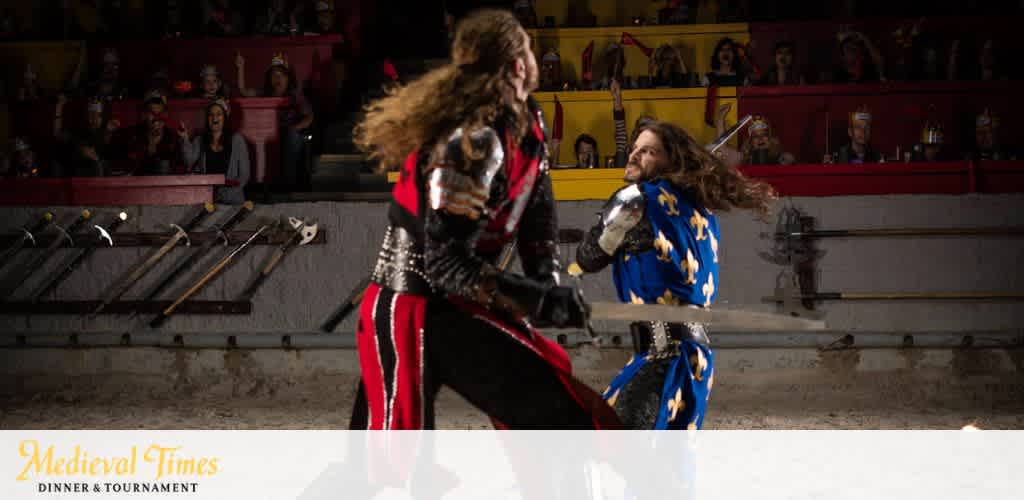 Image shows two knights in a staged combat at a Medieval Times Dinner & Tournament event. One knight, wearing a black and red outfit with a helmet, faces away from the viewer. The other, in a blue tunic with yellow fleur-de-lis, faces toward the viewer, holding a sword ready. An excited crowd watches in the background. The Medieval Times logo appears at the bottom.