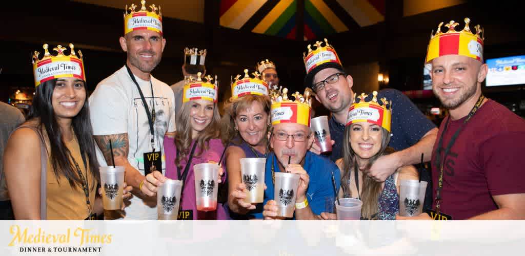 A group of eight happy adults wearing crowns with the Medieval Times logo is posing at a themed event. They are holding up their drinks in a toast-like gesture, smiling, and some have VIP lanyards. The colorful Medieval Times banner is displayed above them.