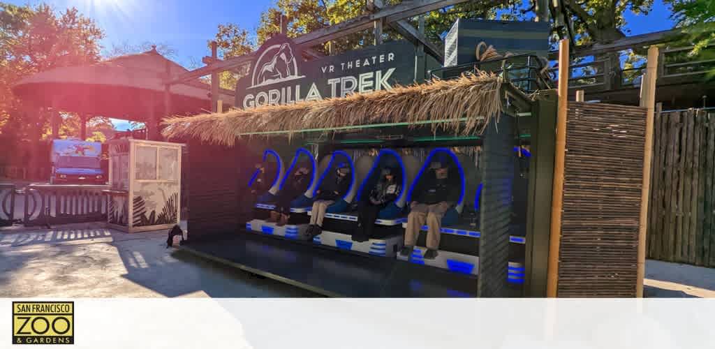 Image shows an outdoor VR theater setup at the San Francisco Zoo & Gardens branded as Gorilla Trek. Guests sit in motion-enabled chairs under a thatched roof, wearing VR headsets. The environment appears sunny and welcoming.