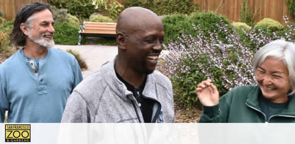 Image of three adults laughing and enjoying a moment together outdoors. Featuring a man with grey facial hair in a blue shirt, a bald man in a grey jacket, and a woman with short grey hair in a green jacket. The background shows purple flowers and greenery. The text 'San Francisco Zoo & Gardens' is visible in the lower left corner of the photo.