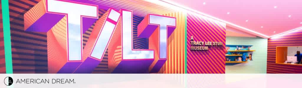 Banner for Tracy Lee Stum Museum at American Dream featuring vibrant, large purple letters 'TLT' on a colorful geometric backdrop. To the right, a reception desk with visitors and staff appears in a brightly lit space with pink and blue walls.