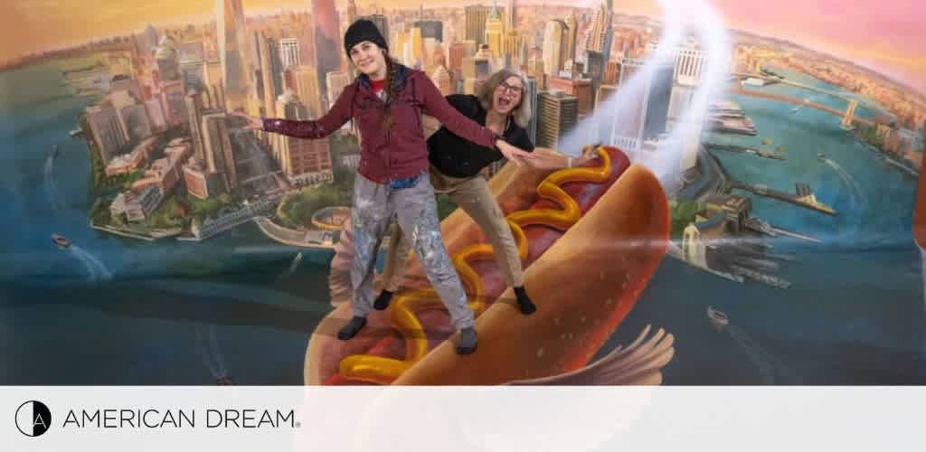 Image shows a vibrant mural at the American Dream mall featuring two individuals playfully posing as if they are surfing atop a giant hot dog flying over a stylized New York City skyline. The backdrop includes iconic buildings, the Hudson River, and a beam of light joining two halves of the scene. The American Dream logo is visible at the bottom.