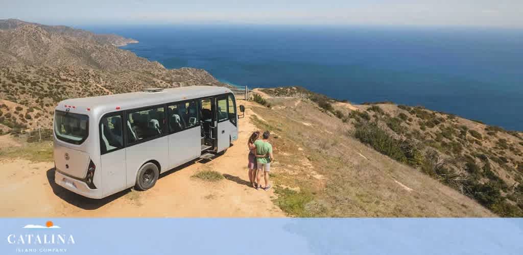 A white shuttle bus is parked on a dirt path alongside a hilly landscape with shrubs and a clear view of the sea. Two visitors, one standing by the open bus door, the other walking on the path, appear to be enjoying the scenic coastal environment on a sunny day. The Catalina Island Company logo is visible in the foreground.