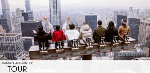 Seven individuals are perched on a railing, overlooking the New York City skyline from a high vantage point. Visible are the silhouettes of various skyscrapers under a cloudy sky. Below them, the words 'Rockefeller Center Tour' suggest the location of this scenic experience.