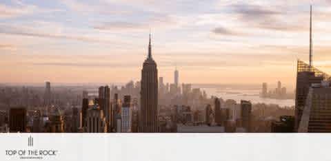 Image displaying a serene sunset view over a city skyline. The Empire State Building dominates the landscape surrounded by other skyscrapers. Soft light bathes the scene, and wispy clouds drape the horizon. Text on the image reads 'Top of the Rock'.