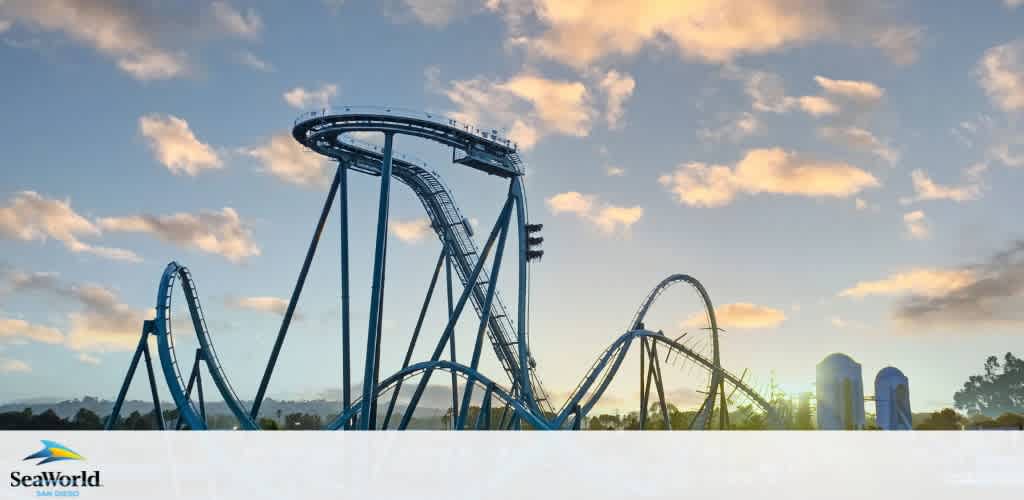 Image displays a towering blue roller coaster with multiple loops against a backdrop of a clear sky peppered with white clouds. The sun sets in the distance, highlighting the silhouette of the attraction. SeaWorld's logo is visible at the bottom left.