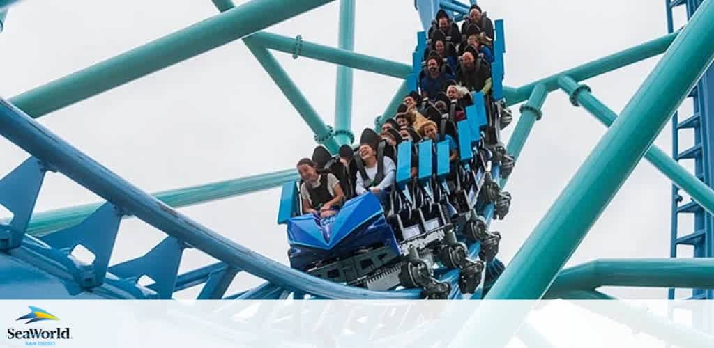 Image shows a group of people on a blue roller coaster at SeaWorld. The coaster is captured mid-drop, exhibiting excitement and enjoyment on the riders' faces. The coaster structure is a tangle of blue steel against a cloudy sky. SeaWorld logo visible at the bottom.