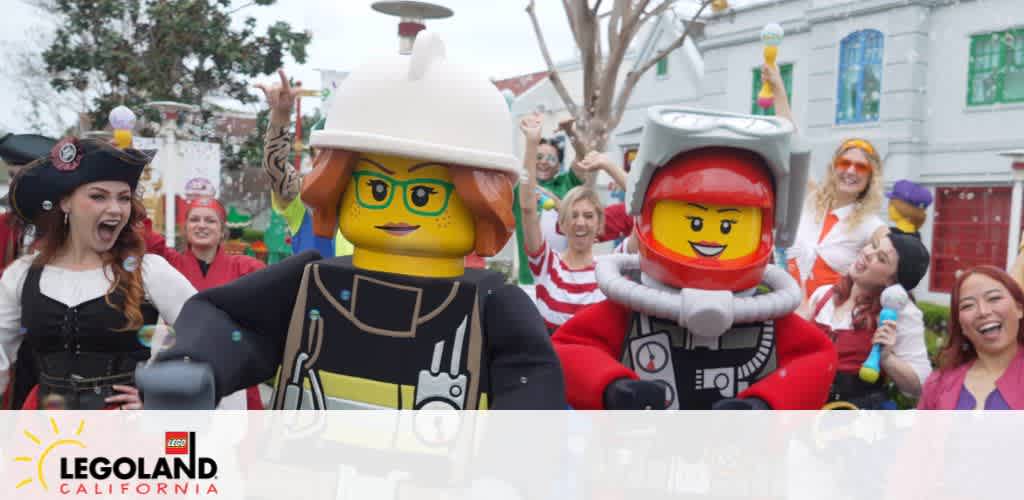 Image displays life-sized Lego figures, one dressed as a firefighter and one as a pilot, with joyful park guests at LEGOLAND California. The visitors, dressed in a variety of costumes, appear to be having an animated celebration. The LEGOLAND California logo is visible at the bottom.