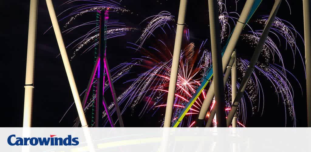 Fireworks light up the night sky above neon-lit roller coaster tracks at an amusement park.