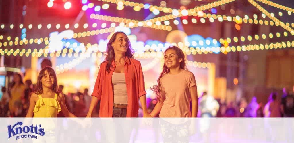 Image of three joyful visitors at Knott's Berry Farm amusement park. They are strolling under a canopy of twinkling lights at dusk. A young girl in a yellow top walks on the left, holding hands with a smiling woman in the center wearing a red cardigan and white top. Another girl in a pink shirt appears on the right, all enjoying the vibrant evening atmosphere. The park’s logo is displayed in the bottom left corner.